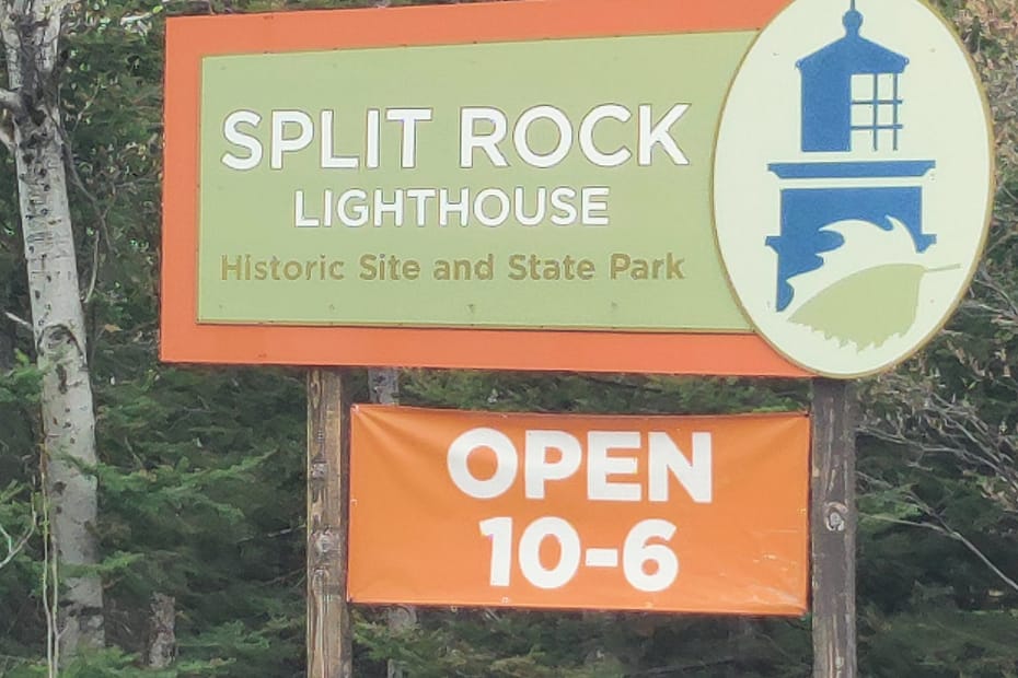 The sign is green with an orange border and white letters reading Split Rock Lighthouse. Below the white words there are green letters stating that it's historic site and park. Next to that is a blue lighthouse design with green and sage waves. Underneath the sign there is another orange sign stating they are open from 10-6 in white letters.
