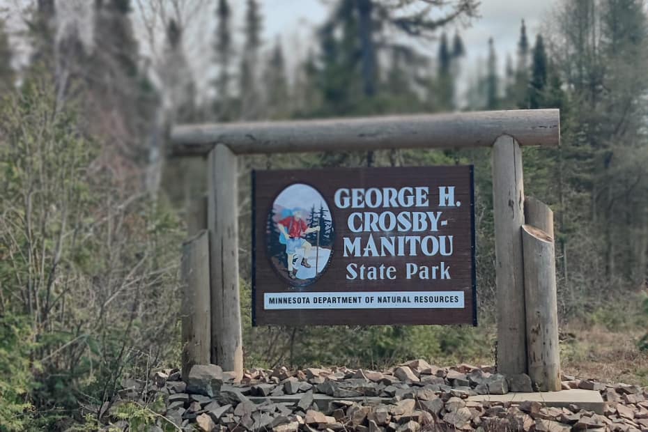 The sign is brown with white lettering that reads GEORGE H CROSBY-MANITOU State Park Minnesota Department of Natural Resources. There is a painting of a hiker on it.