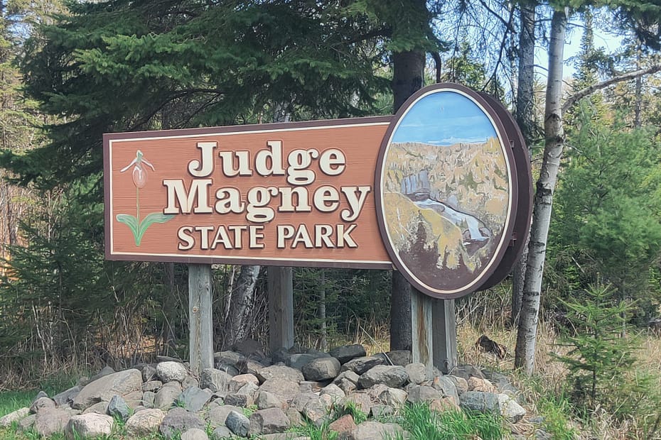 Brown sign with white lettering reading Judge Magney State Park. There is a painting of the river.
