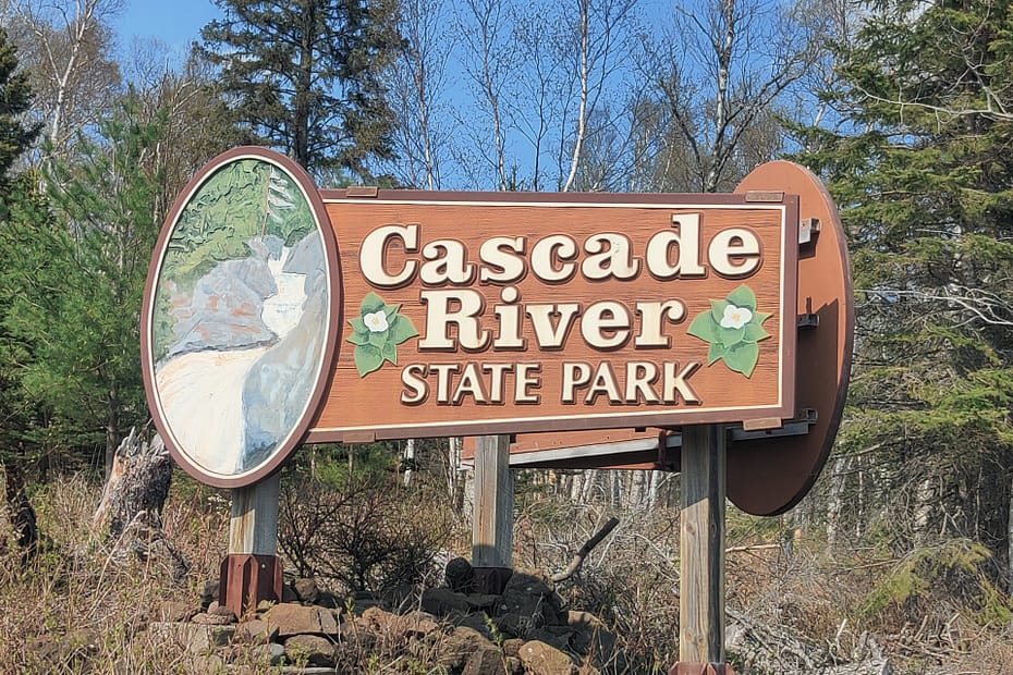 Brown sign with cream-colored lettering that reads Cascade River State Park. There is a painting of the river.
