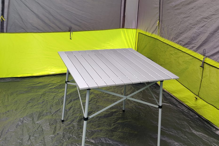 Photo of the Coleman Outdoor Folding Table in Seleana's tent. The table has a slatted aluminum top that attaches to a steel base. The base has crossbars near the top on all four sides.