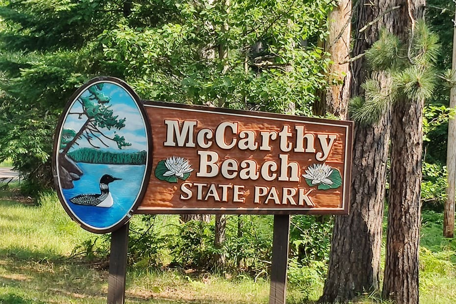 Brown sign with white writing that reads: McCarthy Beach State Park. There is a loon on the lake depicted as well as water lilies.